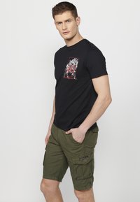 Fekete póló színes grafikai mintával, olajzöld cargo shortsszal párosítva, amely oldalsó zsebekkel és varrási részletekkel rendelkezik.