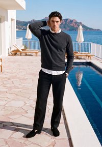 Homme en pull noir et pantalon debout près d'une piscine sur une terrasse en pierre avec des chaises longues et des montagnes visibles de l'autre côté de l'eau.
