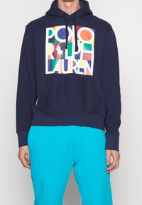 Mörkblå hoodie med en framgrafik med flervägad text "POLO RALPH LAUREN" och en logotyp. Mjukt bomullsmaterial med en känguruficka.