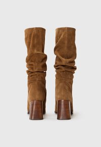 Hnědé suede kozačky do výšky kolen se sklopeným designem a robustními dřevěnými podpatky, s jemnými detaily prošívání.