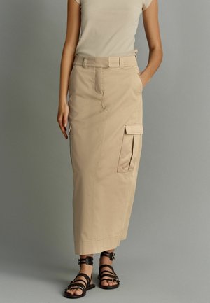 Femme portant une jupe cargo beige, un haut beige et des sandales noires à brides, debout devant un fond gris.