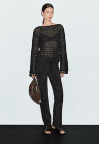 Schwarzes gehäkeltes Strickoberteil mit langen Ärmeln, kombiniert mit schmalen schwarzen Wildlederhosen. Das Model trägt eine braune Schultertasche und schwarze Sandalen mit offenem Zehen.