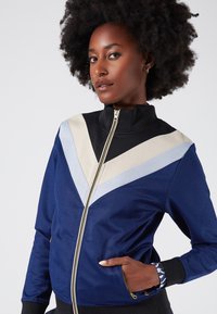 Navy zip-up jacket featuring a high collar, contrasting beige and light blue chevron design, textured fabric, and side pockets.