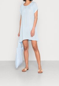 Jasnoniebieska sukienka z krótkim rękawem, plisowana, z detalem w kształcie dziurki. W zestawie z torbą w paski i beżowymi płaskimi sandałami. Gładka tekstura.