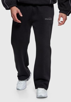 Osoba nosząca czarne, luźne dresy z logo "Dropsize" na udzie oraz białe buty sportowe, spacerująca na jednolitym tle.