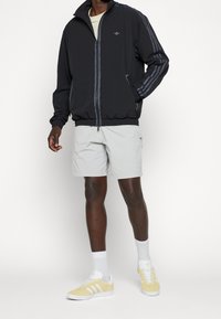 Man med svart zip-up jacka med grå ränder, ljusgrå shorts, vita strumpor och gula och vita sneakers, som står mot en vit bakgrund.