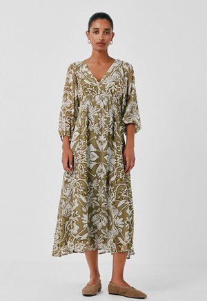 Femme portant une longue robe fleurie vert olive et blanche avec des manches trois-quarts et des chaussures plates beiges, debout devant un fond uni.