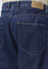 Σκούρο μπλε τζιν ντένιμ από την πίσω όψη που δείχνει δύο πίσω τσέπες, θηλιές για ζώνη και ένα καφέ δερμάτινο λογότυπο με την ένδειξη "SAMSOE SAMSOE."