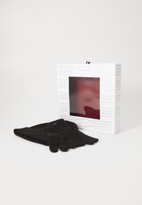 Guantes de lana negros colocados junto a una caja blanca con una ventana transparente, que presenta un acento rojo y el logo de la marca en la parte superior.