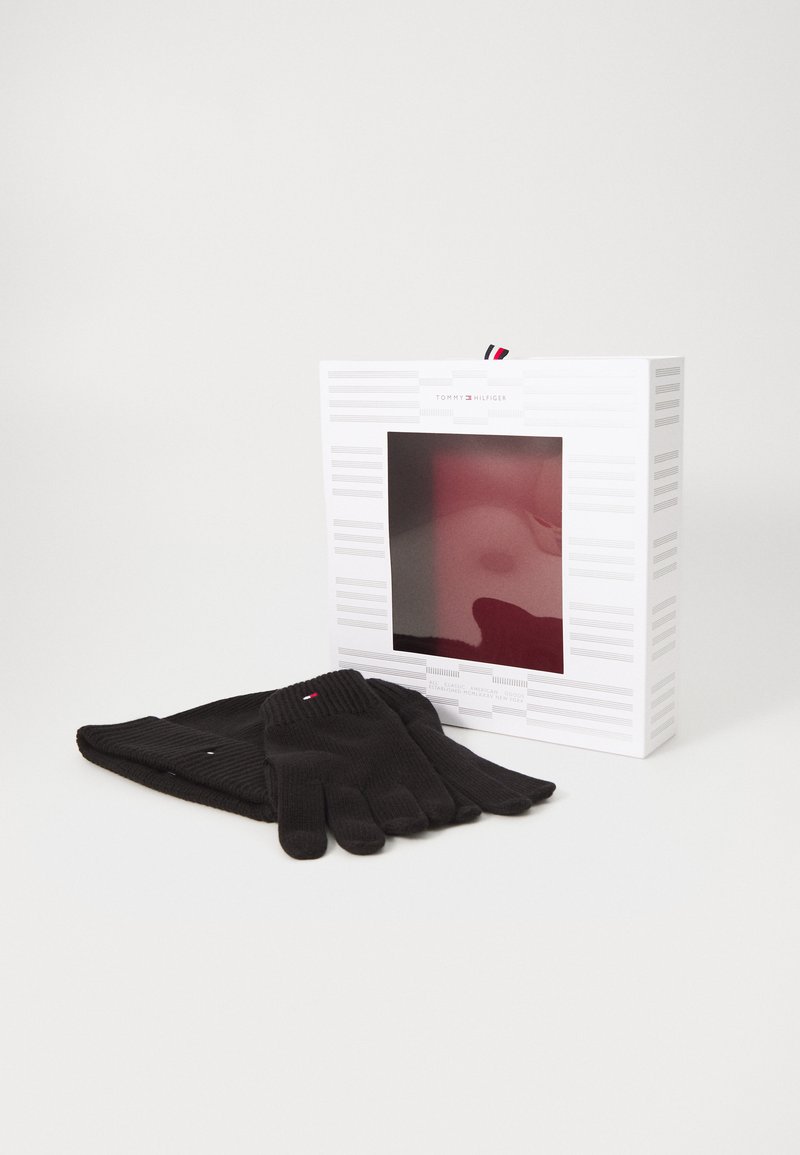 Guantes de lana negros colocados junto a una caja blanca con una ventana transparente, que presenta un acento rojo y el logo de la marca en la parte superior.