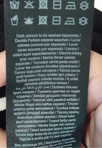 Czarna metka pielęgnacyjna z symbolami prania oraz wielojęzycznym tekstem dotyczącym prania ciemnych kolorów osobno. Zawiera informacje o kurczeniu się.