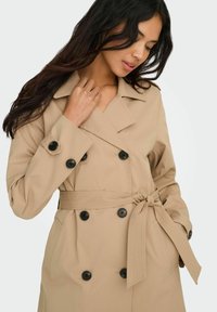 Femme aux longs cheveux bruns portant un trench-coat beige croisé, ceinturé à la taille, avec des boutons noirs visibles.