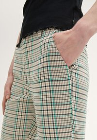 Beige plaid trousers with green, black, and white check patterns. Features side pockets and a relaxed fit. Material has a smooth texture.