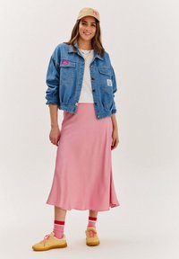 Jeansová bunda s nášivkami, biela košeľa, ružová saténová sukňa, žlté tenisky a viacfarebné ponožky. Hladké textúry a ležérny dizajn.