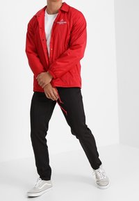 Persoon draagt een rood jasje met knopen over een wit overhemd, zwarte broek met krijtstrepen en witte sneakers, staand met gespreide benen en de handen ineen gevouwen.