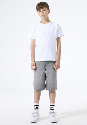 Garçon debout portant un t-shirt blanc, un short gris mi-genou, des baskets blanches et des chaussettes blanches avec des rayures noires et du texte.