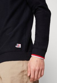Pull en maille bleu marine avec poignets côtelés, présentant un accent rouge et blanc. Comprend une petite étiquette de marque au bas. Tissu texturé.