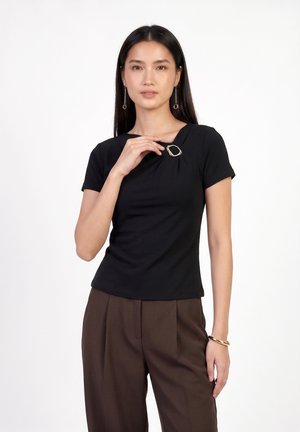 Femme aux longs cheveux foncés portant un haut noir, un pantalon marron, des boucles d'oreilles et un bracelet en or, tenant une broche en or sur son haut.