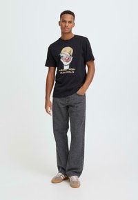 Sort bomuld T-shirt med grafik af en fugl iført kasket og teksten "HEAD HIGH RUN WILD." Kombineret med grå løstsiddende jeans og beige sneakers.