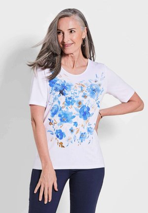 T-shirt blanc à manches courtes avec un imprimé floral bleu et des accents beiges. Encolure dégagée et coupe décontractée. Porté avec un pantalon marine.