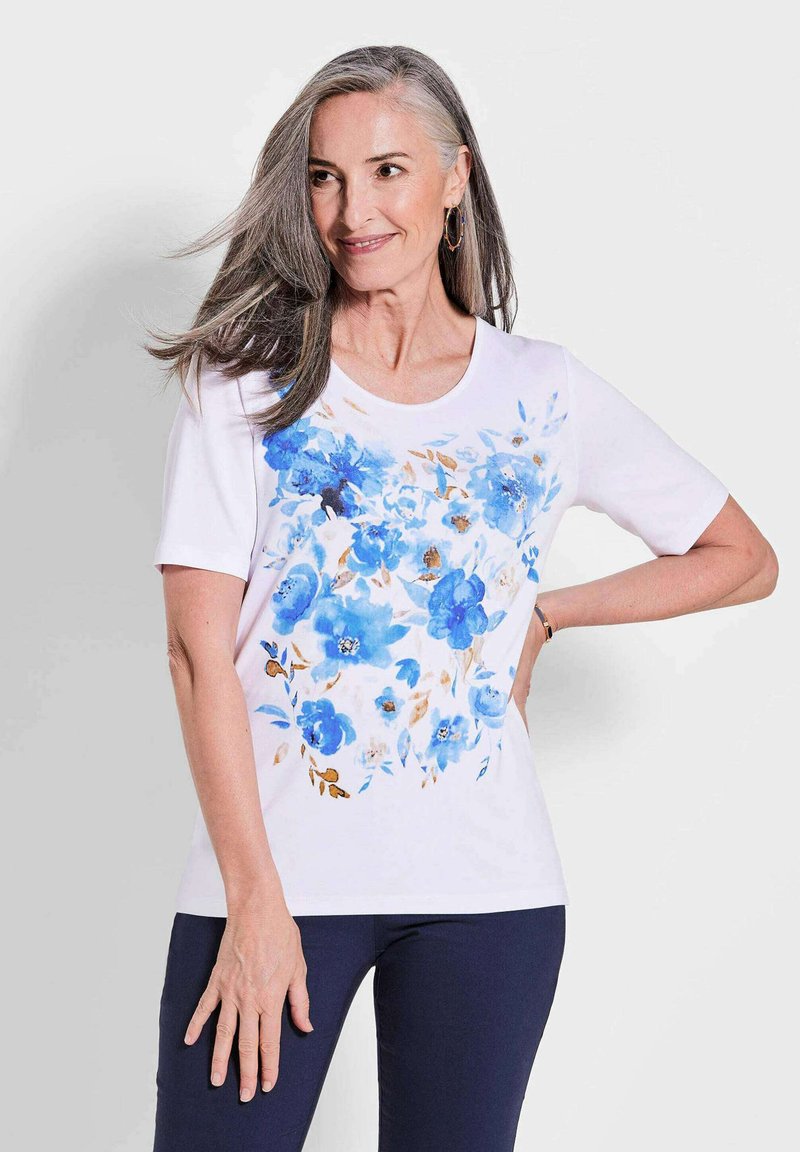 Witte T-shirt met korte mouwen, voorzien van een blauw bloemenpatroon en beige accenten. Ronde halslijn en een comfortabele pasvorm. Gemaakt in combinatie met marineblauwe broeken.