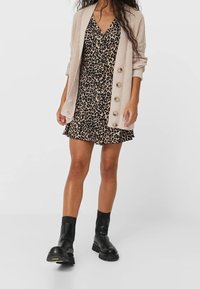 Mujer con un vestido de estampado de leopardo, un cárdigan beige con botones y botas negras hasta el tobillo, de pie frente a un fondo blanco liso.