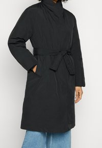 Abrigo acolchado negro con cuello alto, cintura ceñida y bolsillos laterales. Tejido texturizado con un acabado suave y un diseño hasta la rodilla.