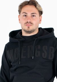 Felpa nera con cappuccio realizzata in tessuto morbido, caratterizzata da un grande logo in rilievo sul petto, coulisse regolabili sul cappuccio e una tasca frontale.