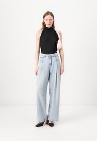 Zwarte geplooide haltertop, lichtblauwe wijde jeans en zwarte hoge hakken; houdt een kleine roze handtas vast. Neutrale achtergrond.