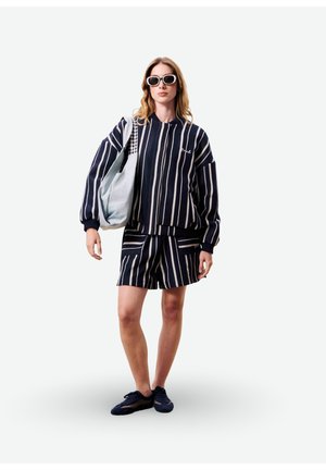 Femme portant une veste à manches longues à rayures verticales bleu marine et blanches et un short assorti, des lunettes de soleil blanches, des chaussures noires, tenant un sac fourre-tout bleu clair.