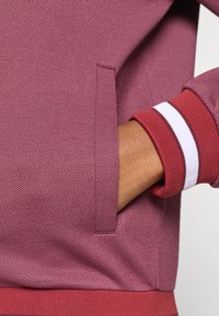 Trumpas rankovės drabužis iš tekstūruoto bordo audinio, turintis šoninę kišenę ir raudonus bei baltus juostelius ant rankovių.