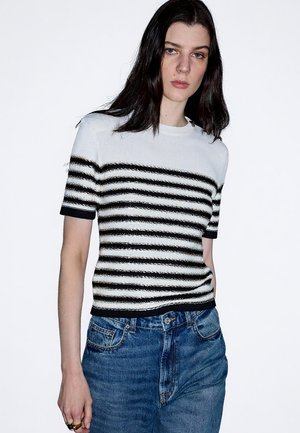 Giovane donna con capelli scuri lunghi che indossa un maglione a maniche corte bianco e nero a righe e jeans blu, in piedi contro uno sfondo bianco uniforme.