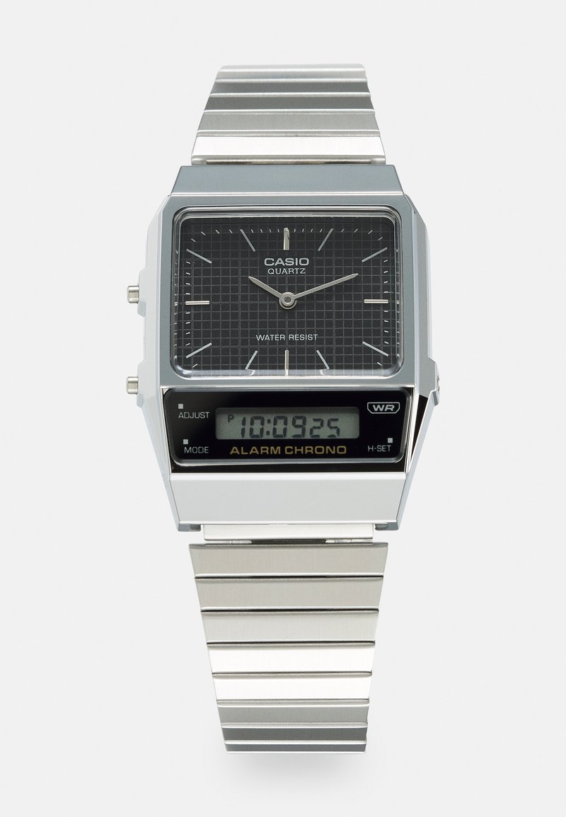 Relógio Casio de quartzo com caixa retangular em prata, mostrador preto com padrão de grelha e display digital. Bracelete metálico com textura escovada. À prova d'água.
