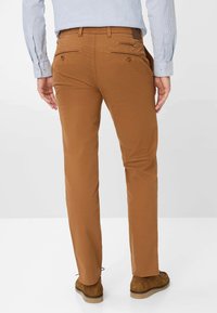 Chinos marroni con una vestibilità slim, dotati di tasche posteriori con zip, tessuto testurizzato e orlo pulito. Abbinati a calzature marroni.
