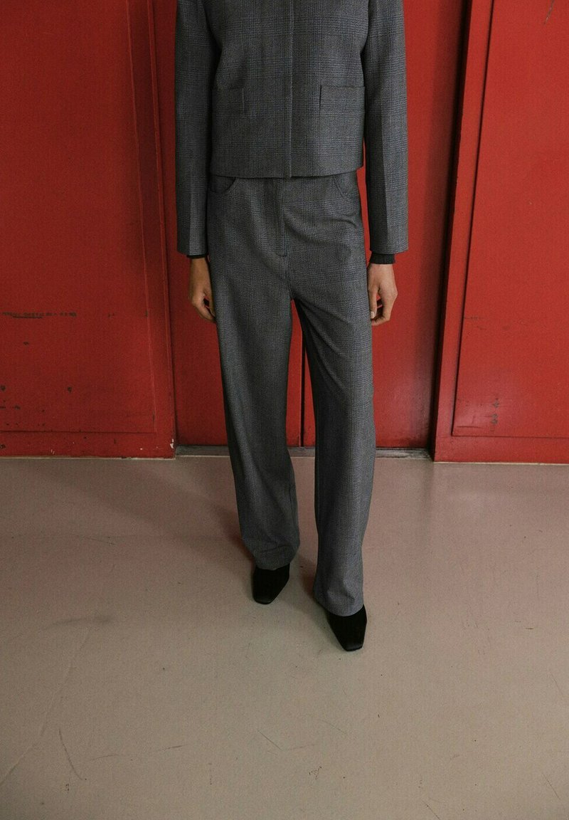 Personne portant un costume en tissu à carreaux gris avec une veste courte et un pantalon à jambes larges, debout devant un mur à panneaux rouges.
