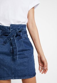 Denimrock mit hohem Bund und Bindegürtel, in Dunkelblau, mit glatter Textur und regulärem Saum. Weißes T-Shirt teilweise sichtbar.