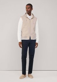 Gilet beige con cappuccio e zip, maglietta bianca a maniche lunghe a costine, pantaloni blu navy e scarpe beige slip-on. Texture liscia, vestibilità casual.
