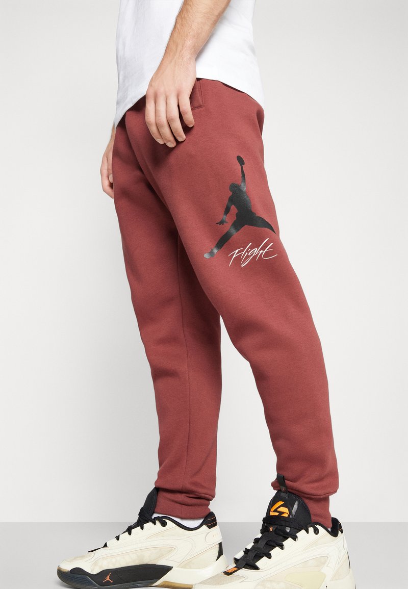 Rote Jogginghose aus weichem Material, mit einem schwarzen Grafiken eines springenden Figures und dem Wort "Flight" in Weiß, kombiniert mit cremefarbenen Sneakern.