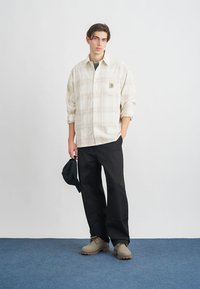 Ανοιχτόχρωμο καρό πουκάμισο με τσέπη στο στήθος, μαύρο παντελόνι, μπεζ ξυλοπάπουτσες και πράσινο καπέλο σε μινιμαλιστικό περιβάλλον με μπλε δάπεδο.