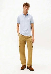Jovem vestido com uma camisola polo azul clara, calças cargo beije e sapatos castanhos, de pé com uma mão no bolso contra um fundo branco.