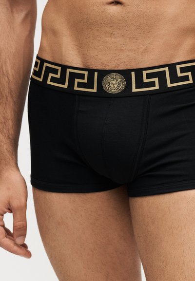 Torso masculin portant un boxer noir avec une ceinture à motif géométrique doré comportant un emblème de tête de Méduse.
