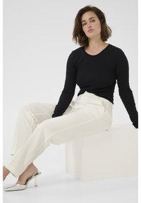 Zwarte longsleeve top gecombineerd met crèmekleurige hoge taille broek en zilveren hakken, zittend op een lichtgekleurde blok.