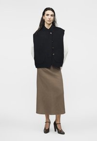 Γυναίκα που φοράει μαύρο αμάνικο γιλέκο με κουμπιά, λευκό μακρυμάνικο τοπ, καφέ μίντι φούστα και μαύρα παπούτσια με ψηλό τακούνι, όρθια.