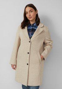 Beige wollen mixjas met een geknipt kraag, voorste knopen, twee zijzakken en een licht oversized pasvorm. Gemaakt over een blauw geruit shirt.