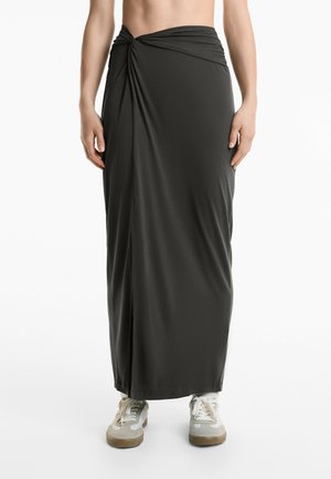 Jupe maxi gris foncé en tissu lisse, avec un design à l'avant tordu et une fente sur le côté, portée avec des baskets claires.