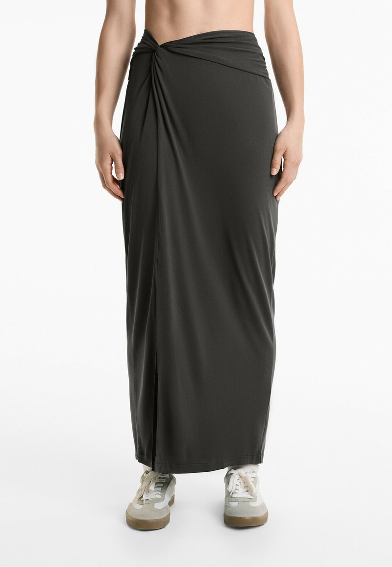 Gonna maxi grigio scuro in tessuto liscio, con un design anteriore attorcigliato e uno spacco laterale, abbinata a sneakers chiare.