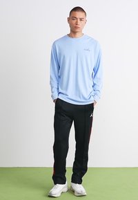 Ung mand står med hænderne i lommen, iført en lyseblå Nike langærmet skjorte, sorte Jordan træningsbukser og hvide sneakers på grøn gulv.