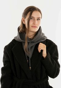 Jonge vrouw met lang haar die een zwarte, gestructureerde trenchcoat over een grijze hoodie draagt, terwijl ze de kraag van de trenchcoat vasthoudt tegen een effen lichte achtergrond.