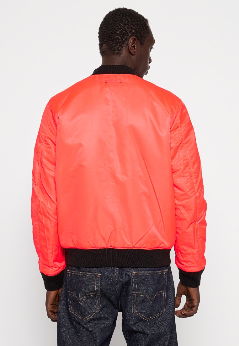 Veste bomber en nylon rouge avec des poignet et un ourlet côtelés noirs, présentant une texture matelassée et une coupe légèrement surdimensionnée.