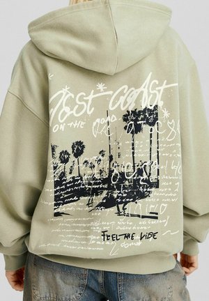 Person iført beige hættetrøje med sort-hvidt palmetræsgrafik, stiliseret håndskrift og teksten "West Coast" og "Feel the Vibe" på ryggen.